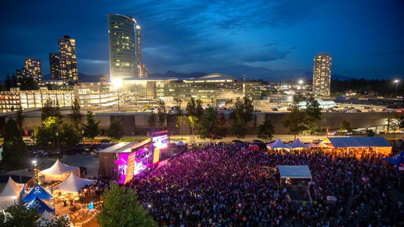 A outdoor music festival at night, with a large crowd and city buildings illuminated in the background.