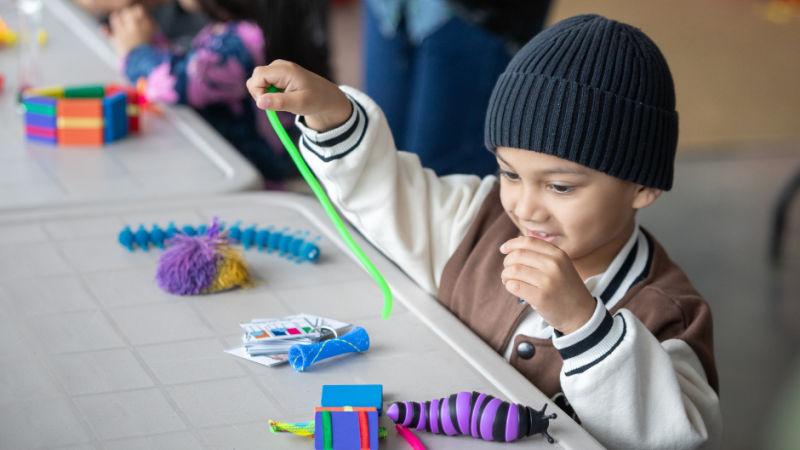 A young child in a beanie cap engaging with sensory play materials at a table during an indoor event.