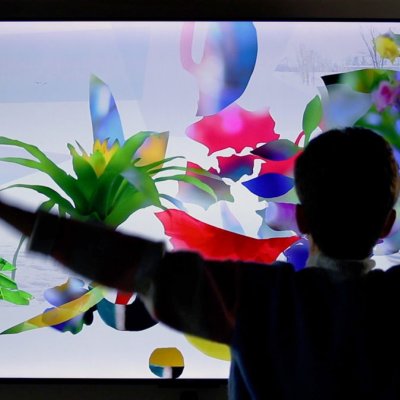 A person with their arms out-wide is in front of an interactive exhibit. Plant shapes of green, red, yellow, blue and purple colours can be seen against a pale blue screen that suggest snow and the outdoors.