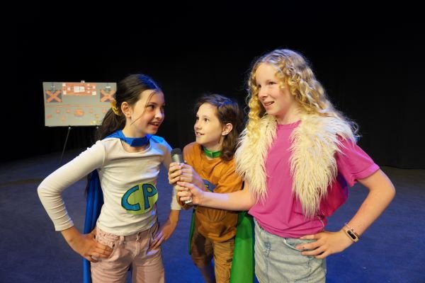 Three students talking into a prop microphone wearing capes. Credit Melissa Regamble